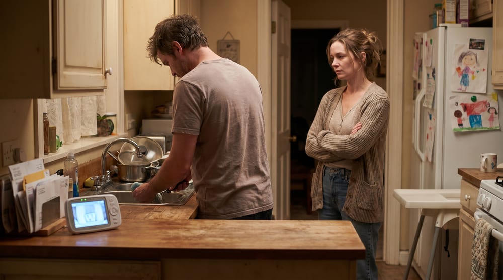 A husband washing dishes with a baby monitor on the counter while his wife stands nearby with her arms crossed, the roommate phase after kids arrive