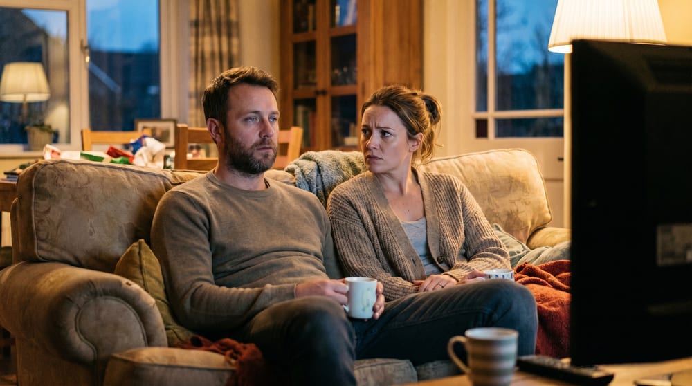 A couple on the couch at night in front of the TV, the husband staring blankly with his mug while his wife watches him with concern