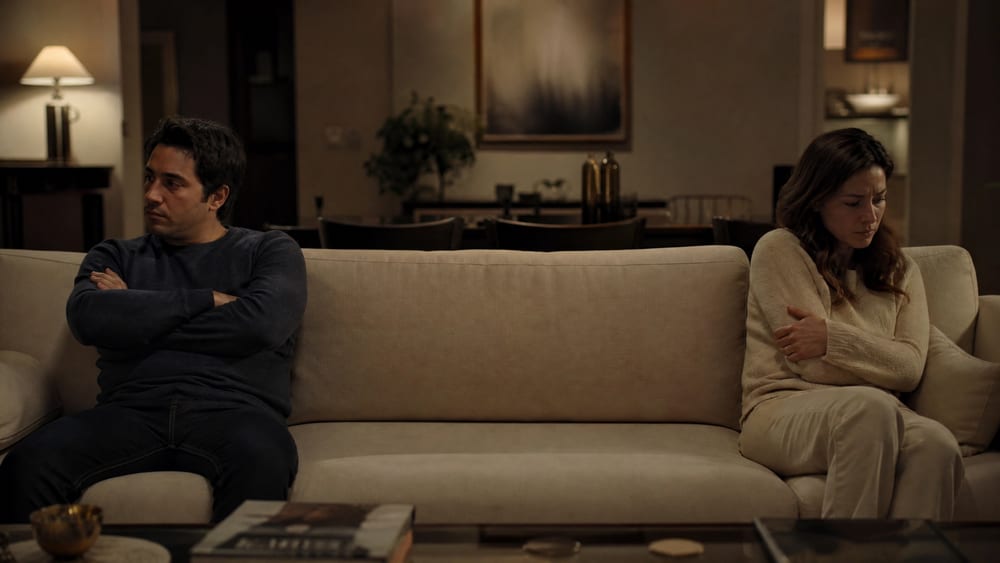 A man sitting with his arms crossed turned away from his wife on the couch, both gone silent after a fight