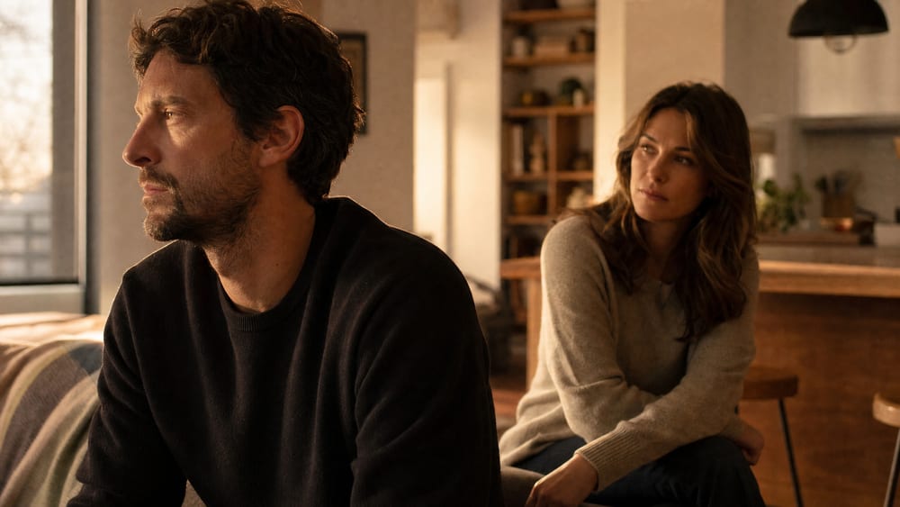 A husband sitting quietly and emotionally withdrawn at home while his wife looks toward him with quiet concern
